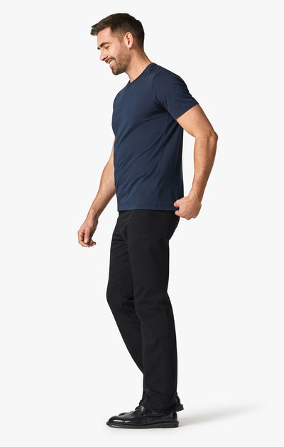 A man stands sideways and smiles, wearing a navy blue t-shirt, black shoes, and 34 Heritage Double Black pant by 34 Heritage featuring a relaxed thigh fit, against a plain white background.