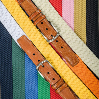 Two brown leather-buckled belts, including a saddle tan style, are placed diagonally over vertically arranged Torino Italian Woven Cotton Elastic Belts in vibrant colors.