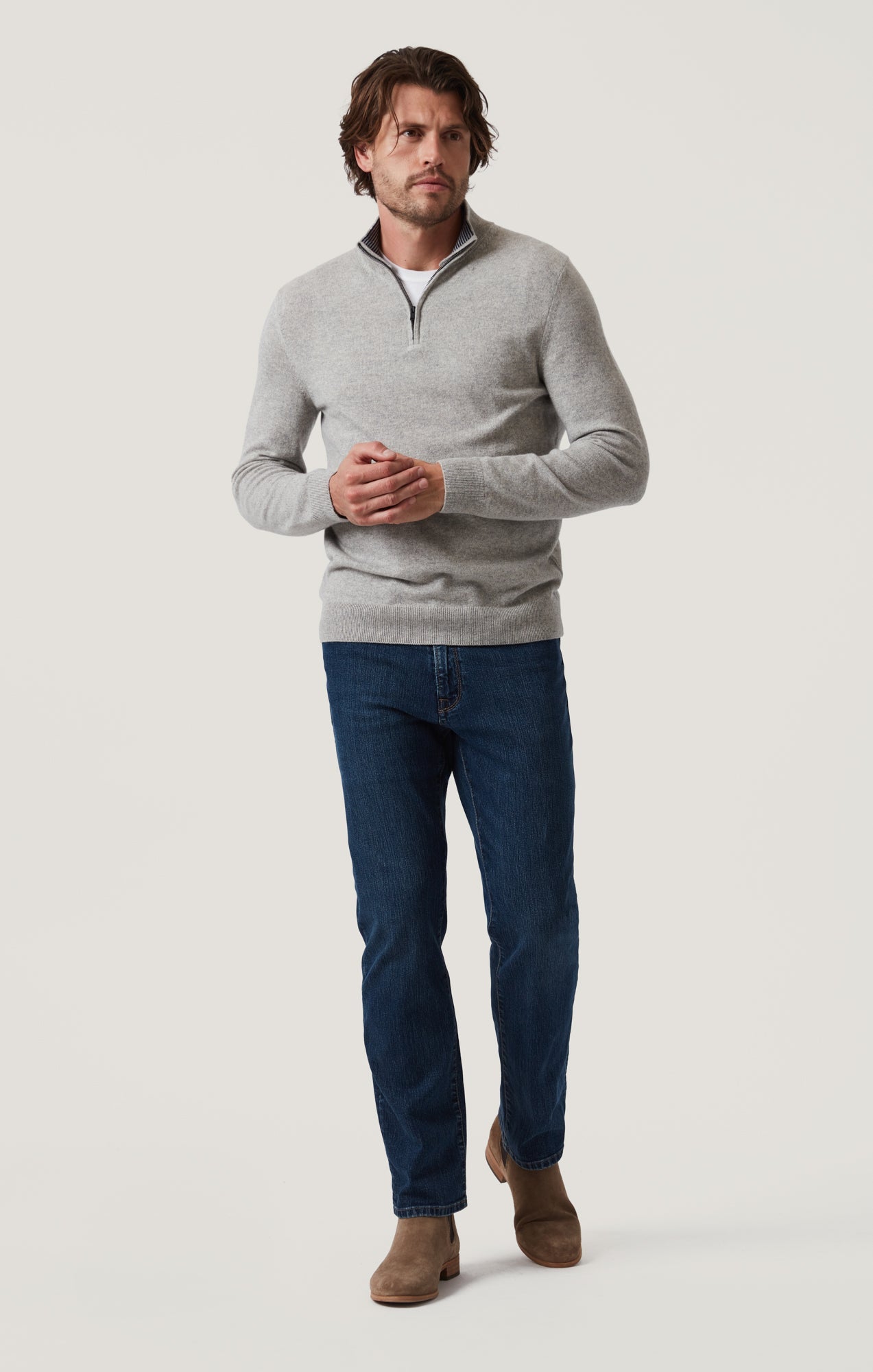A man stands against a plain light background, wearing a light gray quarter-zip sweater, 34 Heritage Mid Comfort higher-rise five-pocket straight leg pants by 34 Heritage, and brown suede boots.