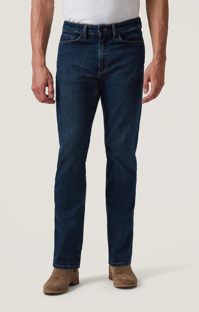 A person wearing a white shirt, 34 Heritage Mid Comfort straight leg fit dark blue jeans by 34 Heritage, and tan suede boots stands against a plain light background.