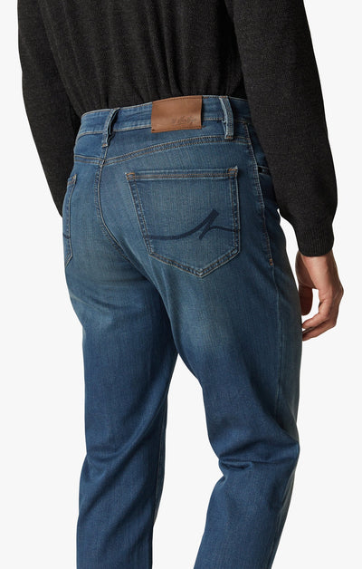 Person shown from the back, wearing 34 Heritage Mid Cashmere high-rise denim blue jeans with a brown leather patch and stitched back pocket design, paired with a black long-sleeve shirt. Jeans feature a soft denim straight leg style by 34 Heritage.