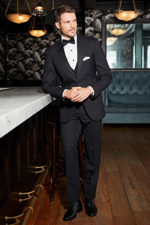 A man in the Michael Kors Black Legacy Performance Tuxedo with a satin notch lapel and bow tie stands by a marble bar in a dim, upscale space with wood floors and vintage decor.