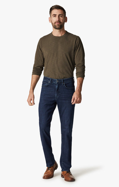 A man faces forward against a white background, wearing an olive green long-sleeve shirt, brown shoes, and 34 Heritage Dark Midnight Brushed Urban modern straight leg jeans by 34 Heritage.