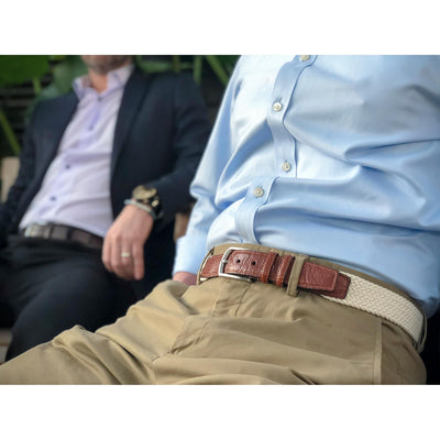 Two men sit together; one wears a light blue shirt, khaki pants, and a Torino Italian Woven Cotton Elastic Belt Big & Tall in saddle tan leather, while the other is dressed in a dark suit with a watch and ring.