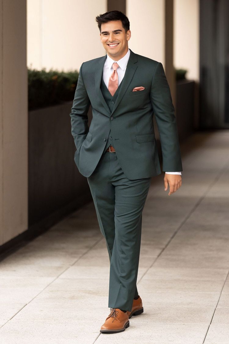 A man in the Ike Behar Hunter Green Luka Suit, featuring a slim fit 100% wool design, pairs it with a peach tie and pocket square as he walks down a hallway smiling with one hand in his pocket.