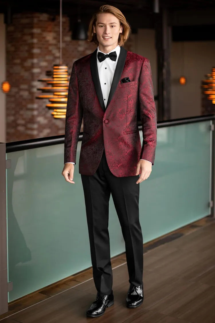 A young man stands indoors in the Mark of Distinction Apple Red Paisley Aries Tuxedo, featuring a patterned red coat with black satin shawl lapel, black pants, a white shirt, and a black bow tie.