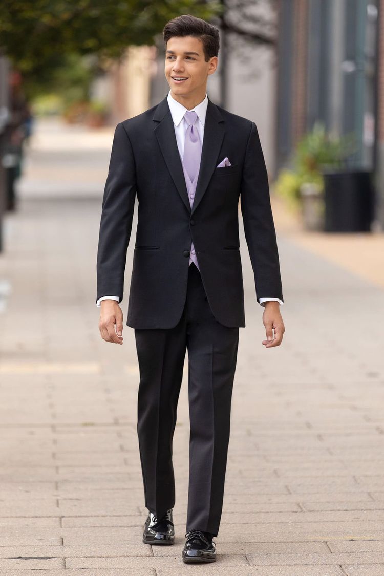 A young man in a modern fit Stephen Geoffrey Black Emerson Tuxedo with a grosgrain notch lapel, styled with a lavender tie and pocket square, walks on a paved urban sidewalk.
