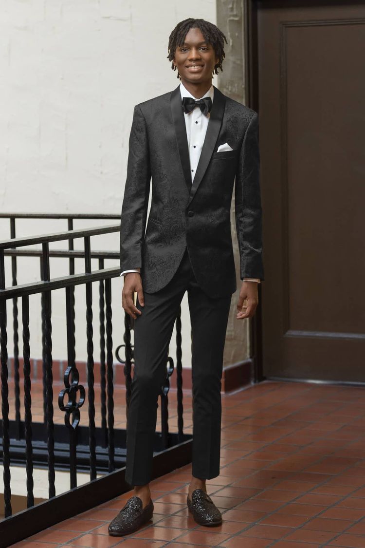 A young man wearing the Mark of Distinction Black Paisley Aries Tuxedo with a black satin shawl lapel, white dress shirt, black bow tie, and loafers stands indoors on a tiled floor beside a railing.