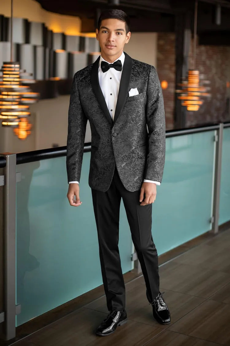 A young man wearing the Mark of Distinction Granite Paisley Aries Tuxedo stands indoors by a glass railing, looking at the camera. He pairs the slim fit tuxedo with a white dress shirt and black bow tie.