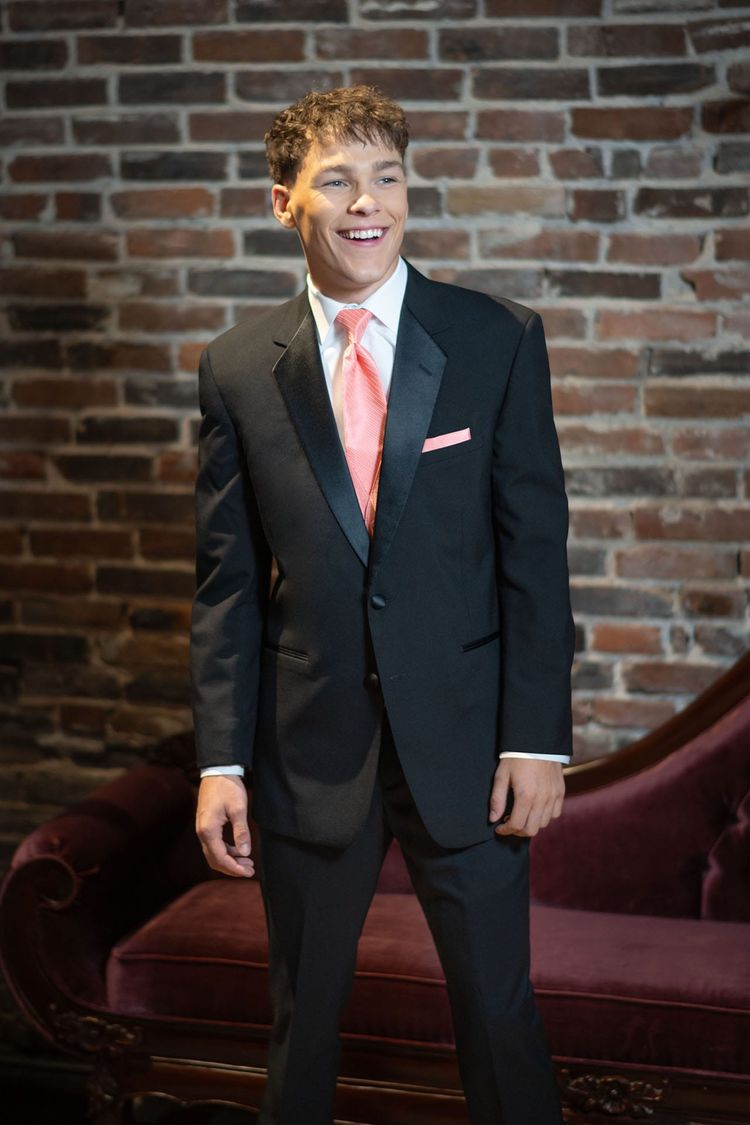 A young man smiles in front of a brick wall and velvet sofa, wearing the Stephen Geoffrey Black Troy Tuxedo in Modern Fit with a satin notch lapel, peach tie, and pocket square. Crafted from 100% worsted wool for a sharp look.