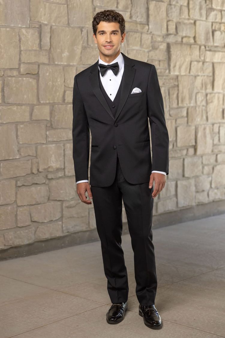 A man wearing the Michael Kors Black Force Performance Stretch Tuxedo with a bow tie stands indoors against a stone wall, looking at the camera.