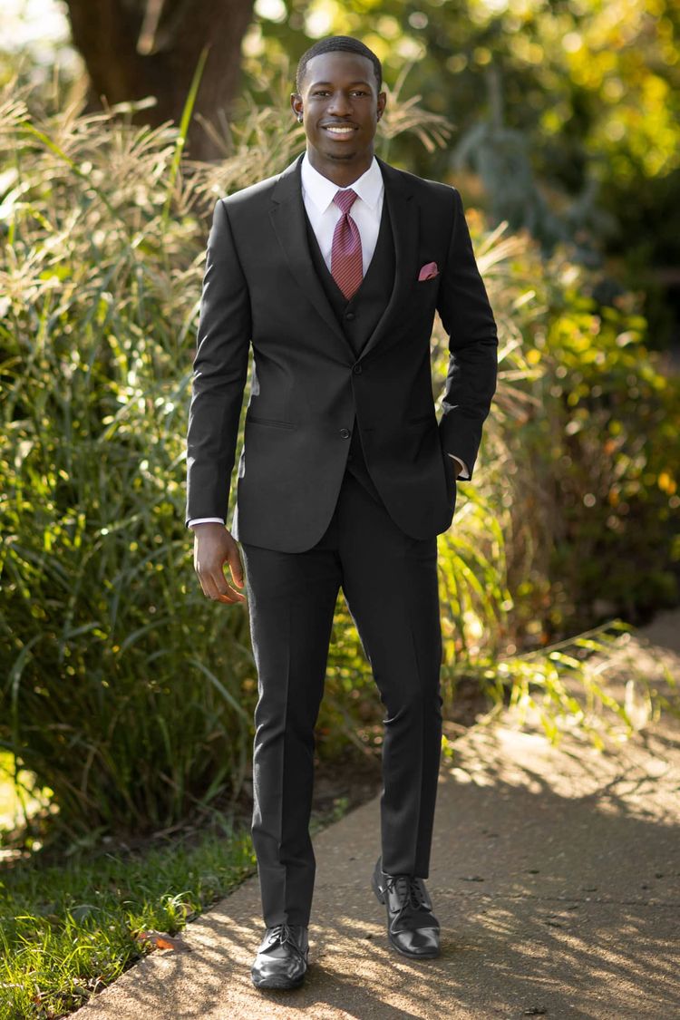 A man in a Michael Kors Black Performance Wedding Suit with a maroon tie and pocket square stands on a sidewalk with greenery—an ideal, stylish option to rent or buy in various men's coat sizes.