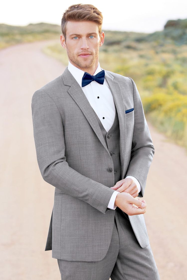 A man in a slim fit Heather Grey Clayton Suit by Allure Men with a navy bow tie stands on a dirt road, surrounded by greenery and yellow flowers—ideal for renting or buying timeless style.