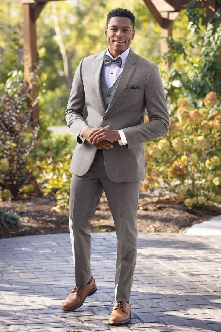 A man wears the Michael Kors Medium Grey Performance Wedding Suit with a bow tie and brown dress shoes, standing on a paved path outdoors amid greenery and wooden structures—ideal inspiration to rent or buy.