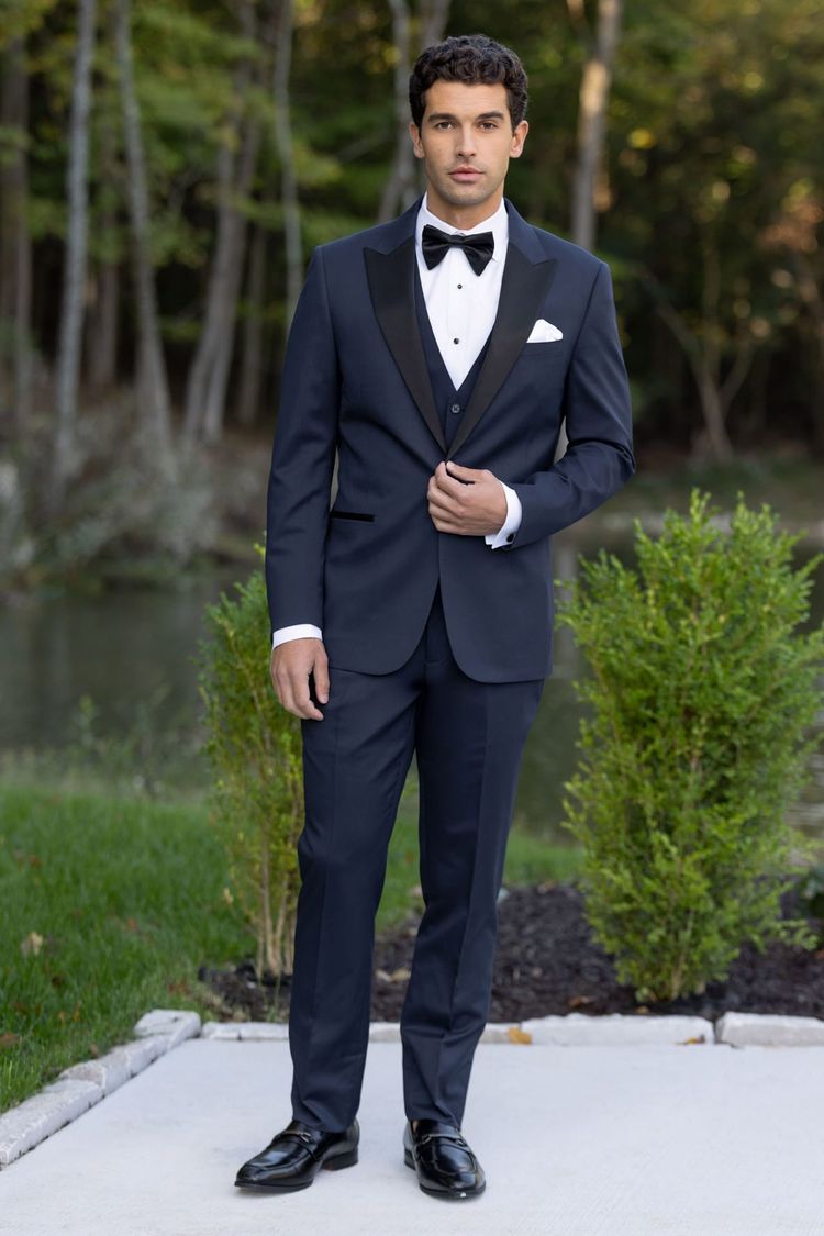 A man in a Michael Kors Navy Performance Stretch Tuxedo with a black satin lapel and black bow tie stands outdoors on a sidewalk, greenery and trees in the background.