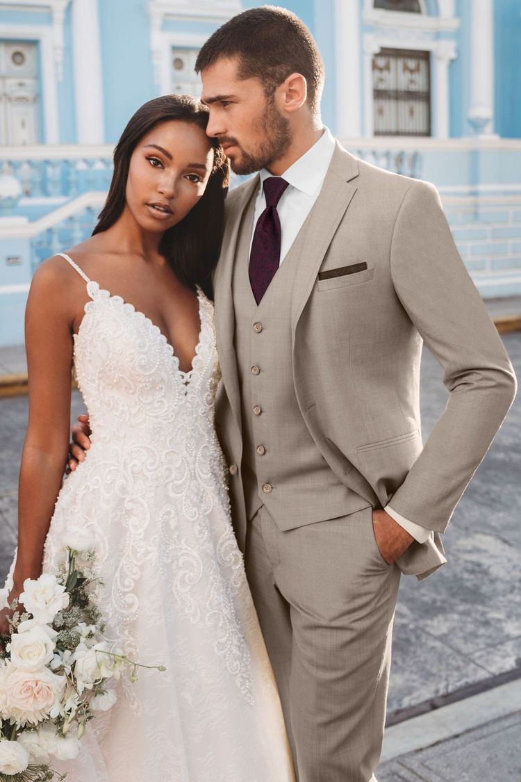 A bride in a white lace wedding dress holds a bouquet beside a groom wearing the Sand Brunswick Suit by Allure Men with a burgundy tie, posing outdoors in front of a blue building.