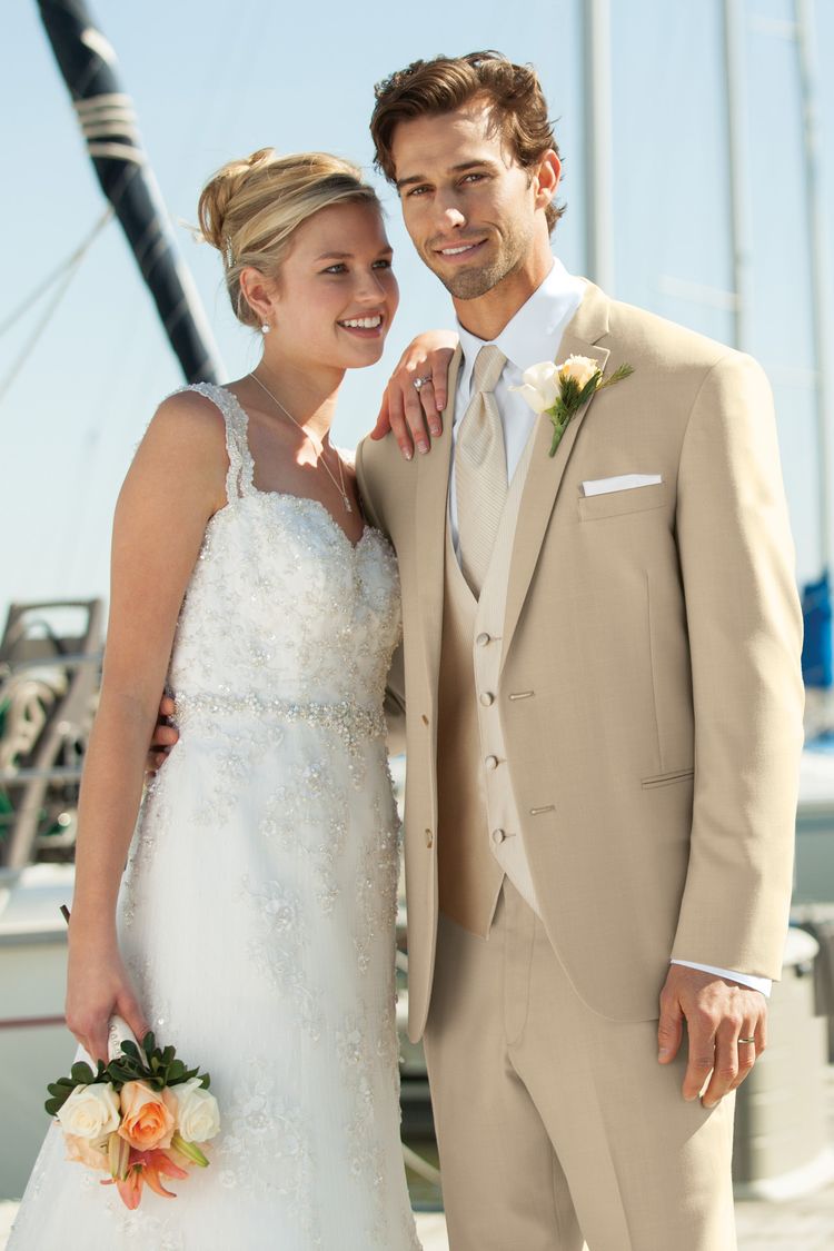 A bride in a white dress and a groom wearing the Lord West Tan Havana Suit stand outdoors, smiling, with boats and masts visible in the background.