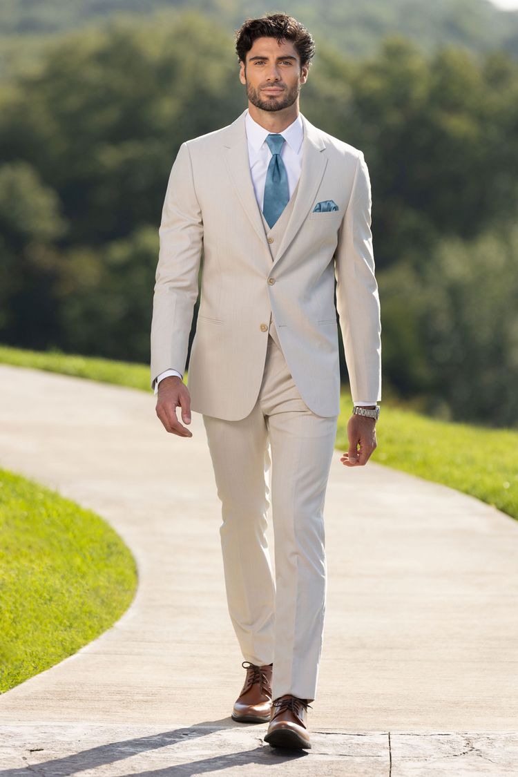 A man in a Michael Kors Tan Performance Wedding Suit walks outdoors amid greenery, styled with a blue tie, white shirt, and brown shoes.