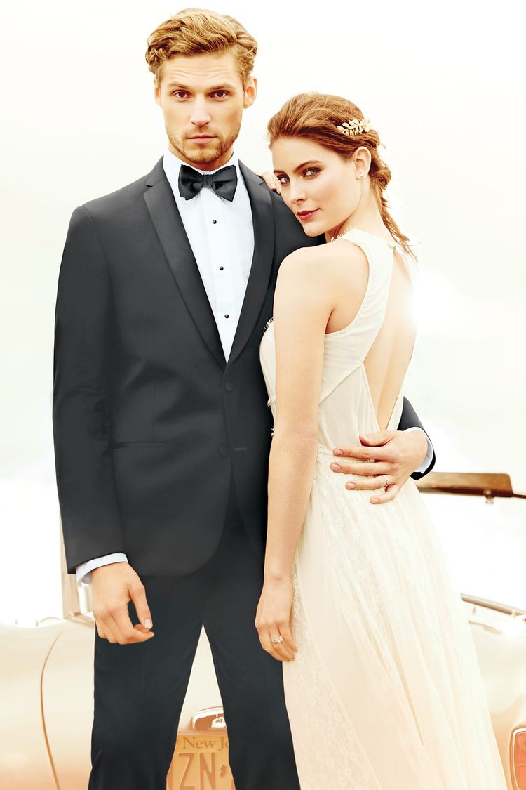 A man wears the Michael Kors Black Berkeley Tuxedo in a modern fit Super 130’s wool, while a woman in a sleeveless white gown poses with him before a beige vintage car for a formal photo.