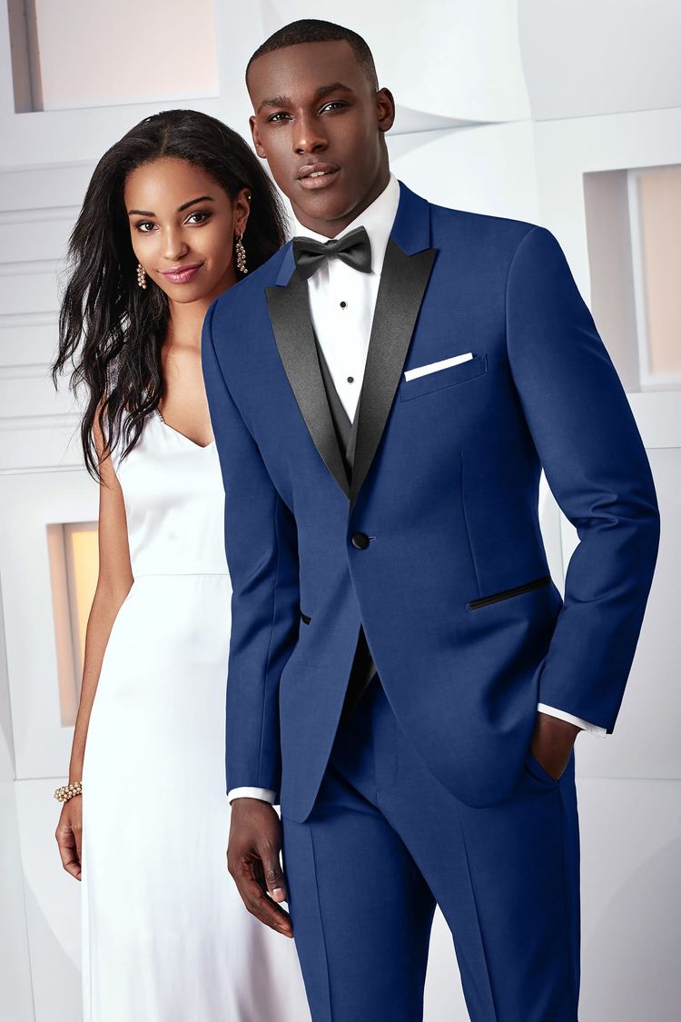 A man wears the Ike Behar Cobalt Blue Tribeca Tuxedo, featuring a slim fit and black satin lapel, as he stands next to a woman in a white sleeveless dress against a light indoor background.