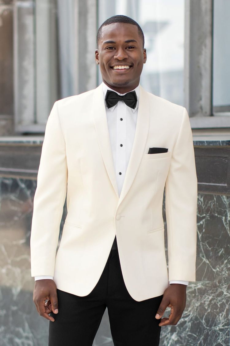 A man stands smiling in front of a marble wall and window, dressed in the Stephen Geoffrey Ivory Classic Shawl Tuxedo—featuring a modern fit coat, self shawl lapel, white jacket, black bow tie, and black pants—available for rent or purchase.