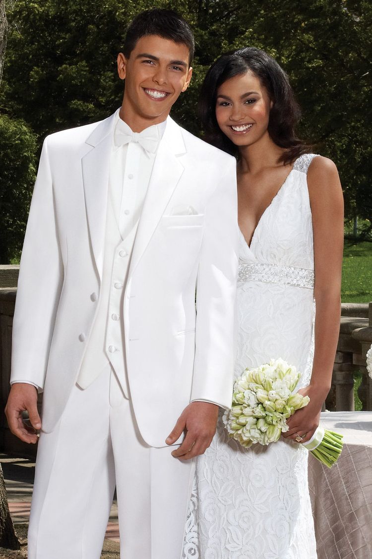 A man wears the Stephen Geoffrey White Troy Tuxedo and stands outdoors with a woman in a white wedding dress, both smiling at the camera as she holds a bouquet of white flowers.