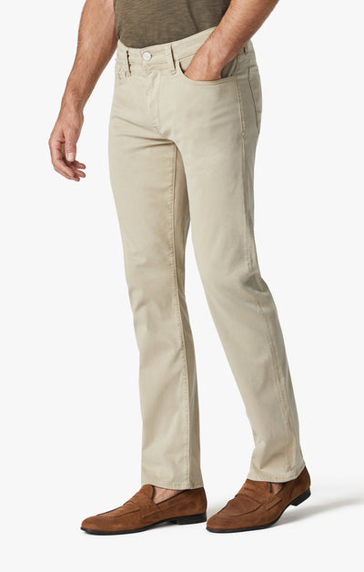 A man wearing 34 Heritage Aluminum Twill pants with a brown belt, brown loafers, and a greenish-brown shirt stands against a plain white background, one hand in his pocket.