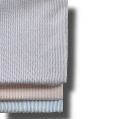 Three folded Wilkes & Riley Twill Stripe Dress Shirts in light gray, pink, and blue are stacked on a white surface, casting a shadow to the right.