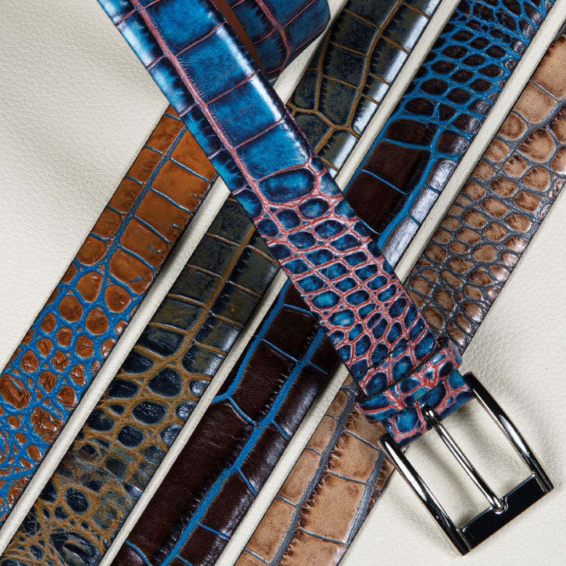 Five luxury Torino Faux Crocodile Embossed Calf Belts in blue, brown, and tan, styled overlapping on a light textured surface.