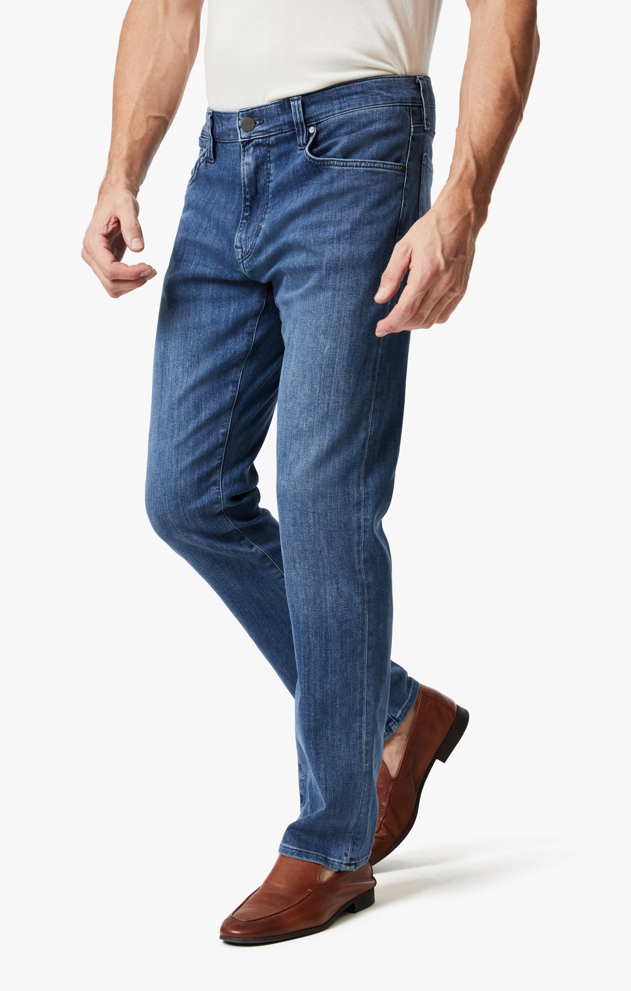 A man wearing 34 Heritage Mid Urban dark-wash jeans by 34 Heritage, a white shirt, and brown dress shoes stands against a plain white background.