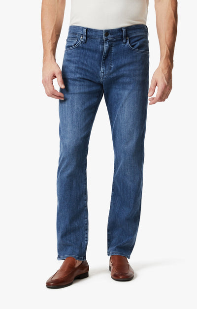 A man stands against a plain white background wearing 34 Heritage Mid Urban dark-wash straight-leg jeans by 34 Heritage, a white shirt, and brown dress shoes.