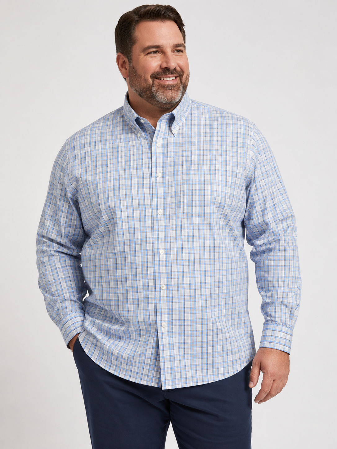 A man is wearing the Tailor & Mason T55-126T Long Sleeve Check Shirt, featuring a blue and grey plaid design in non-iron cotton, paired with dark blue pants. He stands smiling against a plain light background.