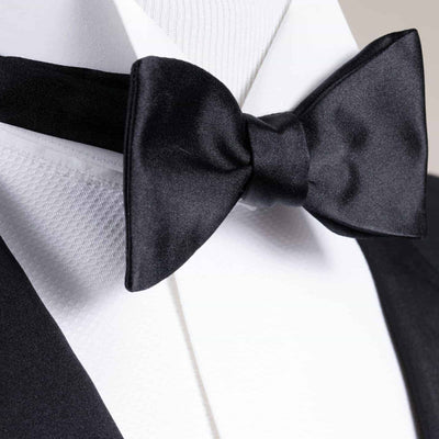 Close-up of the IKE Pre-Tied Silk Bow Tie by Ike Behar, a sleek black silk accessory, styled with a white dress shirt and black tuxedo jacket.