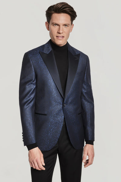 A man wears the Jack Victor Navy Paisley Dinner Jacket from Ted's Clothiers, featuring black lapels, over a black turtleneck sweater and black pants, standing against a plain light background.
