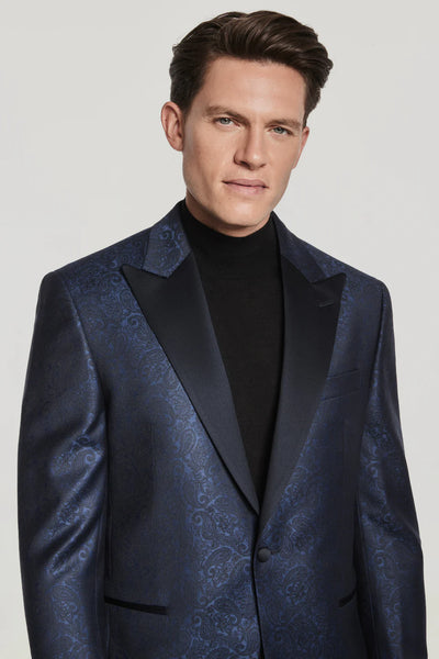 A man wearing the Jack Victor Navy Paisley Dinner Jacket from Ted's Clothiers with black satin lapels and a black turtleneck stands against a plain light background.