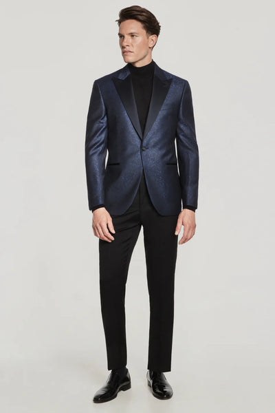 A man stands against a plain light background wearing a Jack Victor Navy Paisley Dinner Jacket from Ted's Clothiers, paired with a black turtleneck, black trousers, and black shoes.