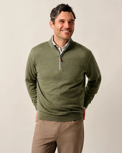 A man smiles with hands behind his back, wearing a Johnnie-O Sweater SULLY 1/4 Zip in olive green over a collared shirt and light brown pants against a plain background.