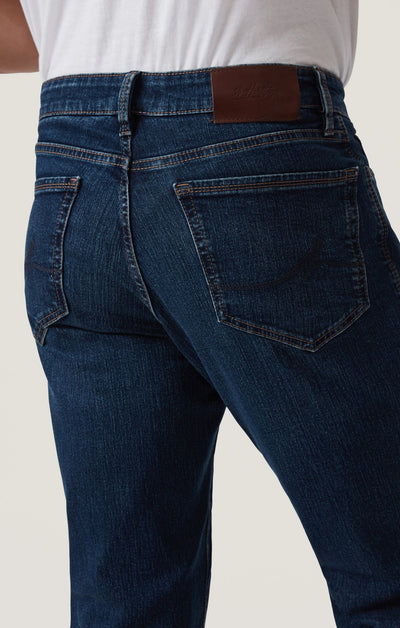 A person is shown from the back wearing 34 Heritage Mid Comfort dark blue denim straight leg jeans and a white shirt, highlighting the higher-rise waistband, rear pockets, and signature brown leather patch by 34 Heritage.