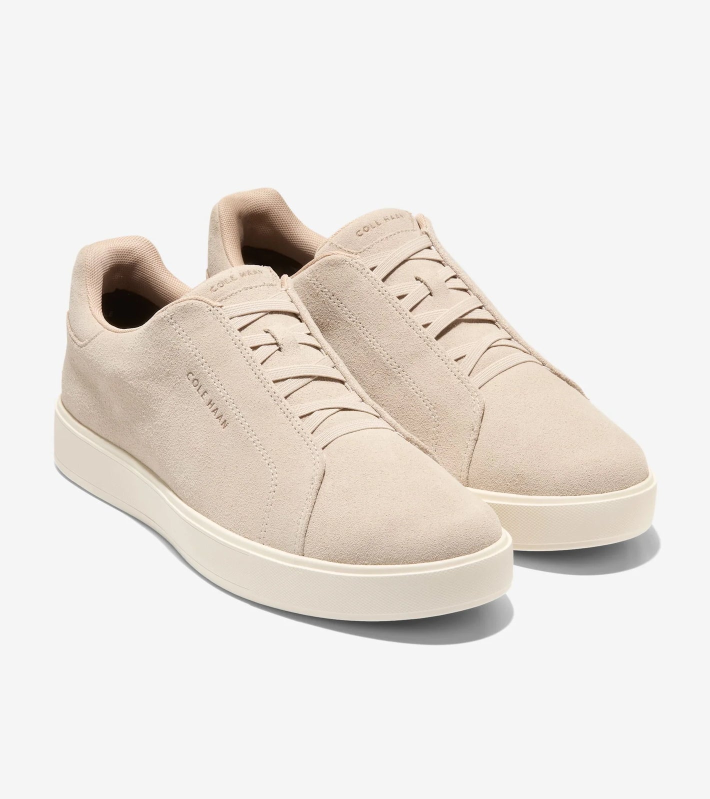 A pair of Cole Haan GrandPro Luxe Slip On Sneakers in beige suede with white rubber soles and minimal stitching, featuring FlowerFoam™ cushioning, shown on a plain white background.