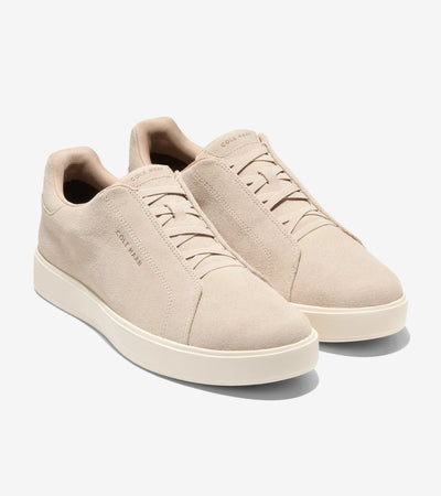 A pair of Cole Haan GrandPro Luxe Slip On Sneakers in beige suede with white rubber soles and minimal stitching, featuring FlowerFoam™ cushioning, shown on a plain white background.