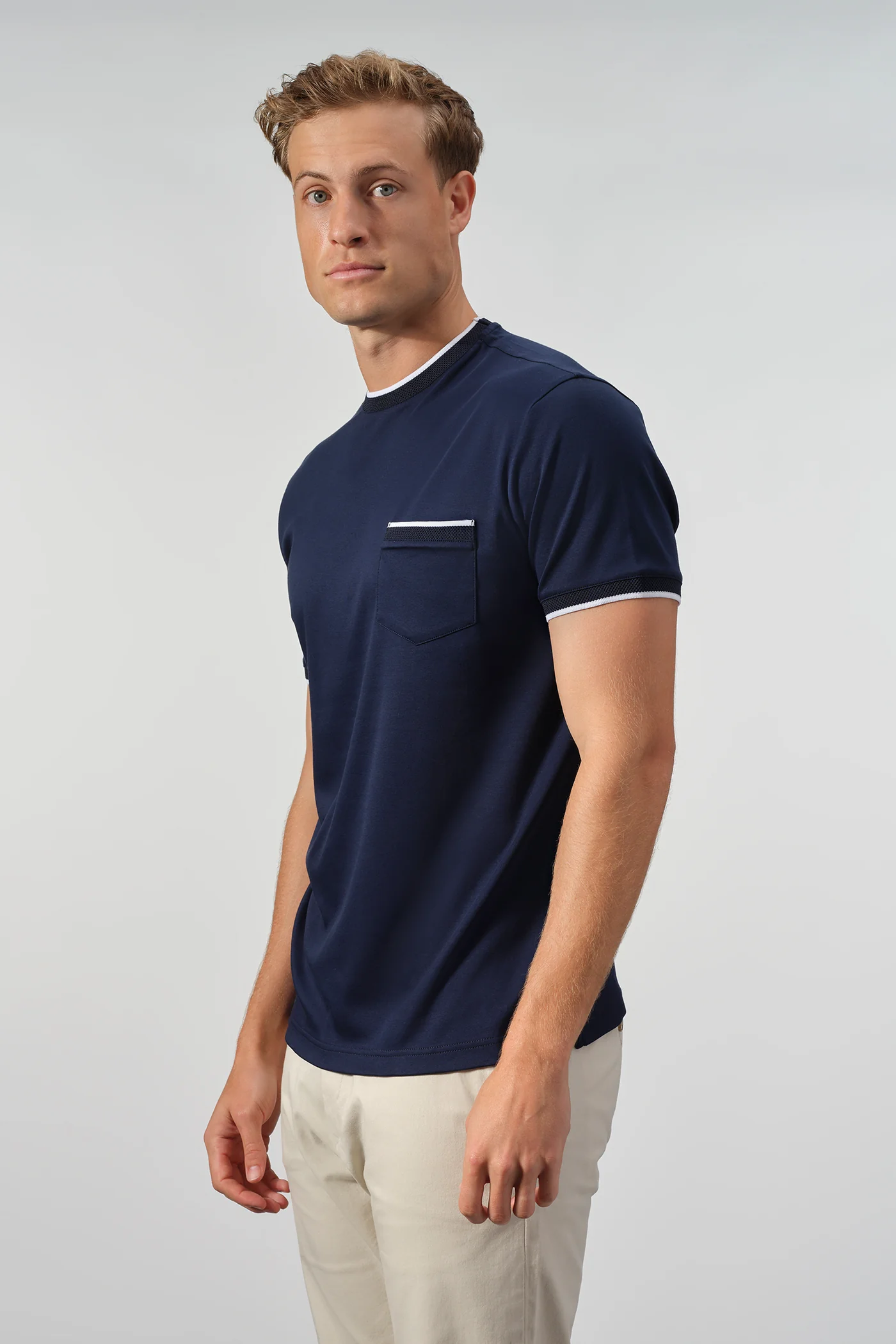 A man stands against a plain light background wearing the Garnet Setatouch Pocket Tee 2617011CC by Garnet, featuring navy blue fabric with white trim on the collar and pocket, paired with light-colored pants.
