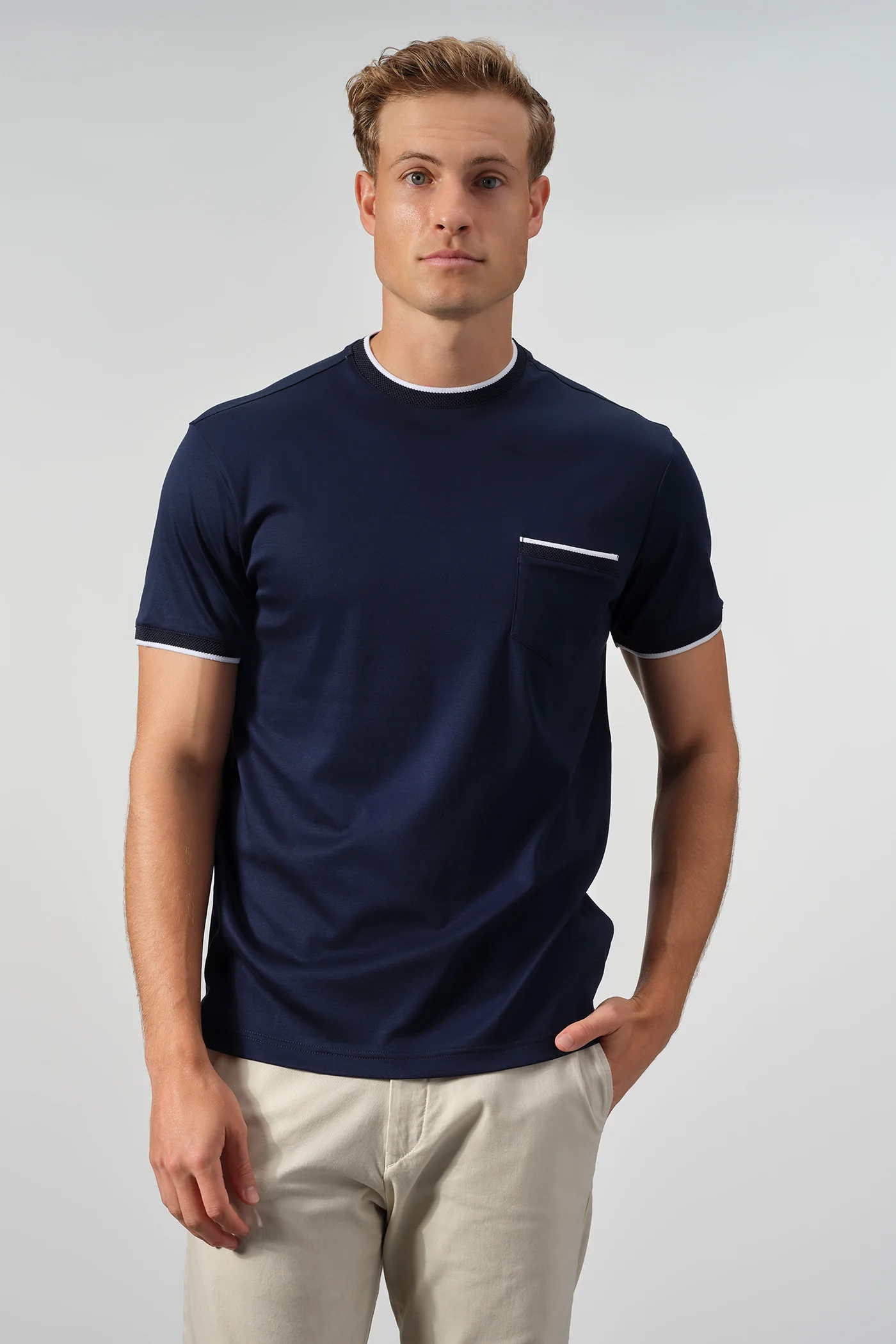 A man stands against a plain background wearing the Garnet Setatouch Pocket Tee 2617011CC by Garnet, a navy blue short-sleeve t-shirt with white trim and chest pocket, paired with light beige pants.