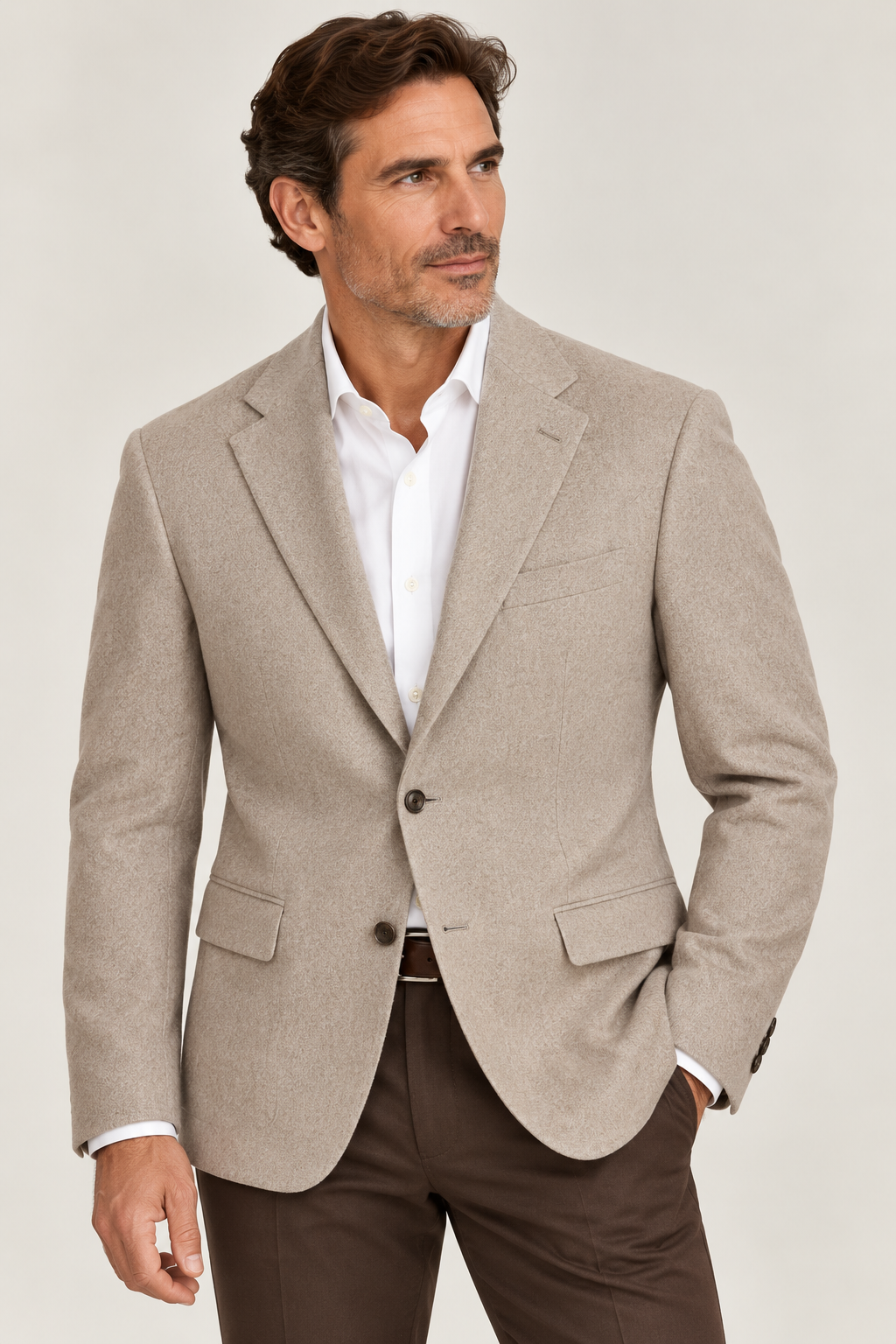 A man wearing the Giles & Jasper Cashmere Sport Coat by Giles & Jasper in beige, styled over a white dress shirt and brown pants, stands against a plain light background, gazing slightly to the side.