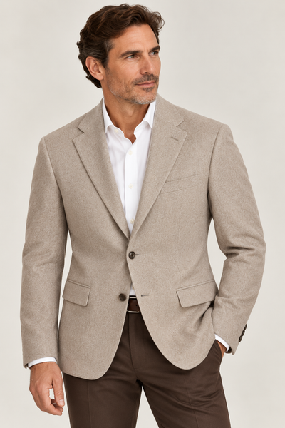 A man wearing the Giles & Jasper Cashmere Sport Coat by Giles & Jasper in beige, styled over a white dress shirt and brown pants, stands against a plain light background, gazing slightly to the side.
