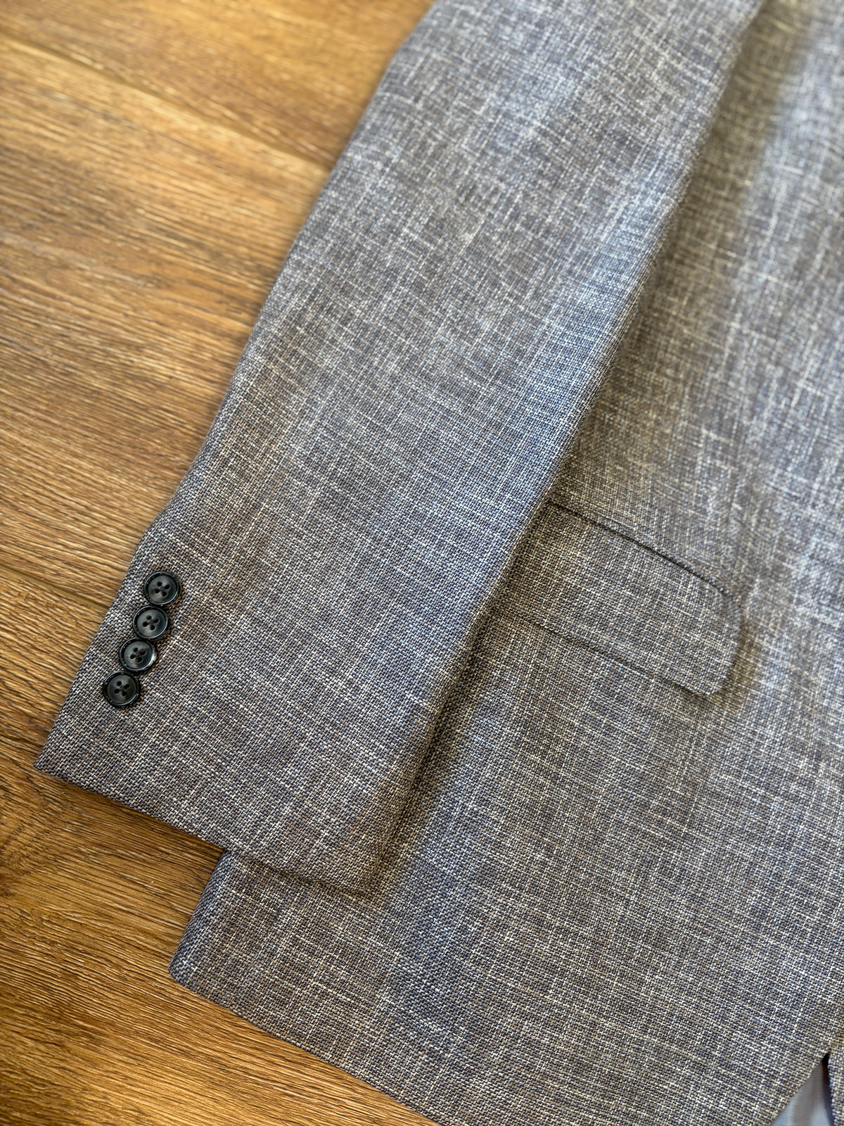 Close-up of the sleeve of the Hart Schaffner Marx Grey and Brown New York Sport Coat 3900010, featuring a gray textured fabric and four black buttons, resting on a wooden surface.