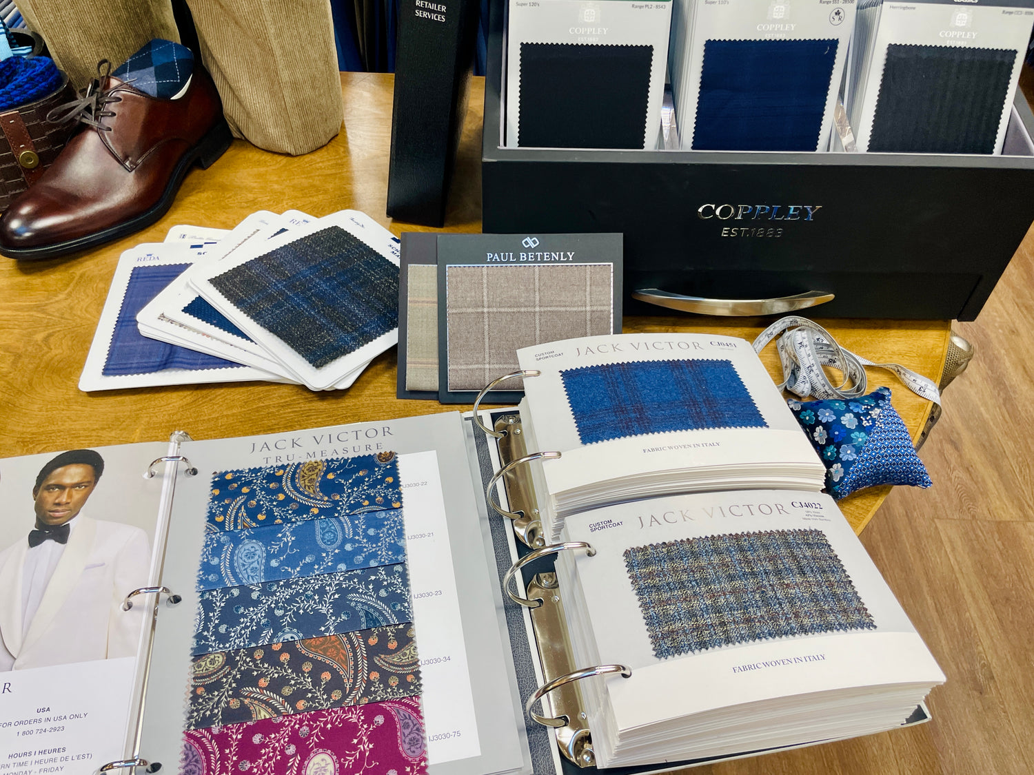 A display of fabric swatches, sample books, and accessories on a wooden table, including suit fabrics, dress shoes, and handkerchiefs.