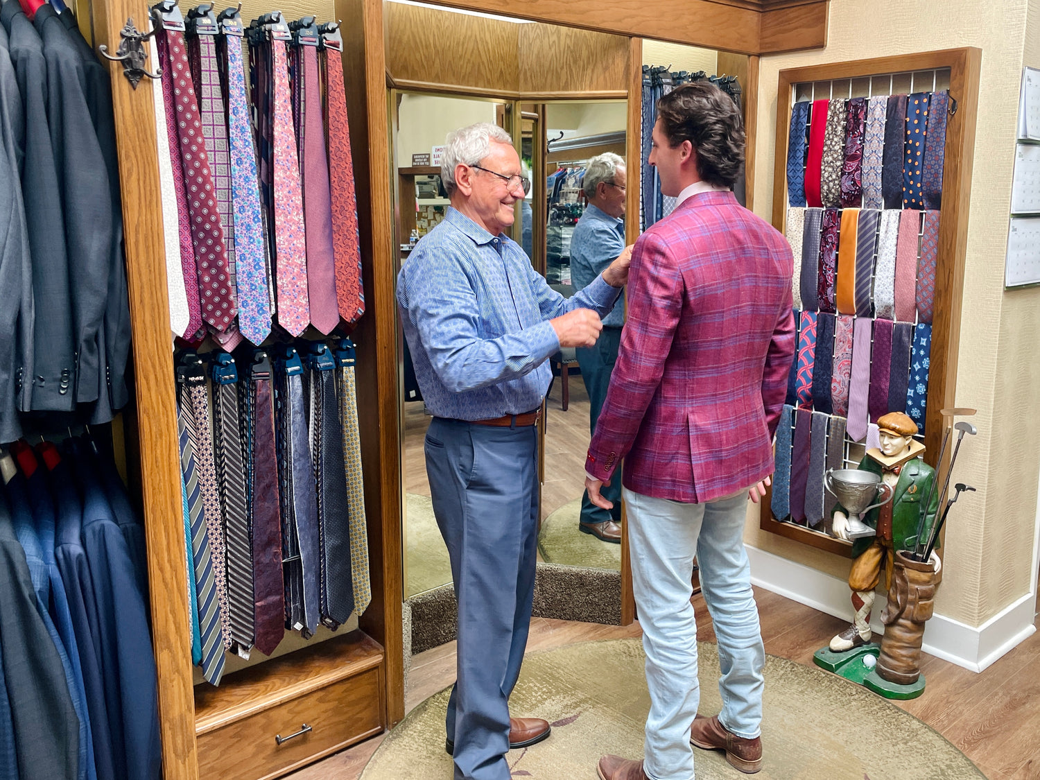 Two men stand in a clothing store; one helps the other adjust a blazer. Racks of neckties and suits are visible, and a wooden statue stands near the mirror.