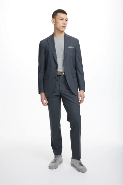 A man stands against a plain white background wearing the Jack Victor Wool Stretch Suit in dark gray by Jack Victor, paired with a light gray t-shirt and gray sneakers.