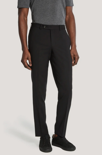 A man stands with his arms relaxed at his sides, wearing a grey short-sleeve shirt, black Jack Victor Riviera Traveler Pants, and black sneakers.