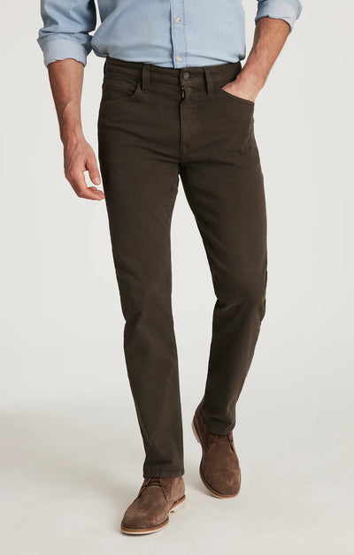 A man wears 34 Heritage Courage Diagonal Pants in coffee, paired with a light blue button-down shirt and brown suede shoes, standing with one hand in his pocket.